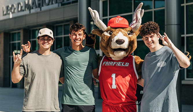 Three young men smile while posing with a cheerful Lucas the Stag mascot, showcasing a fun and lively atmosphere.